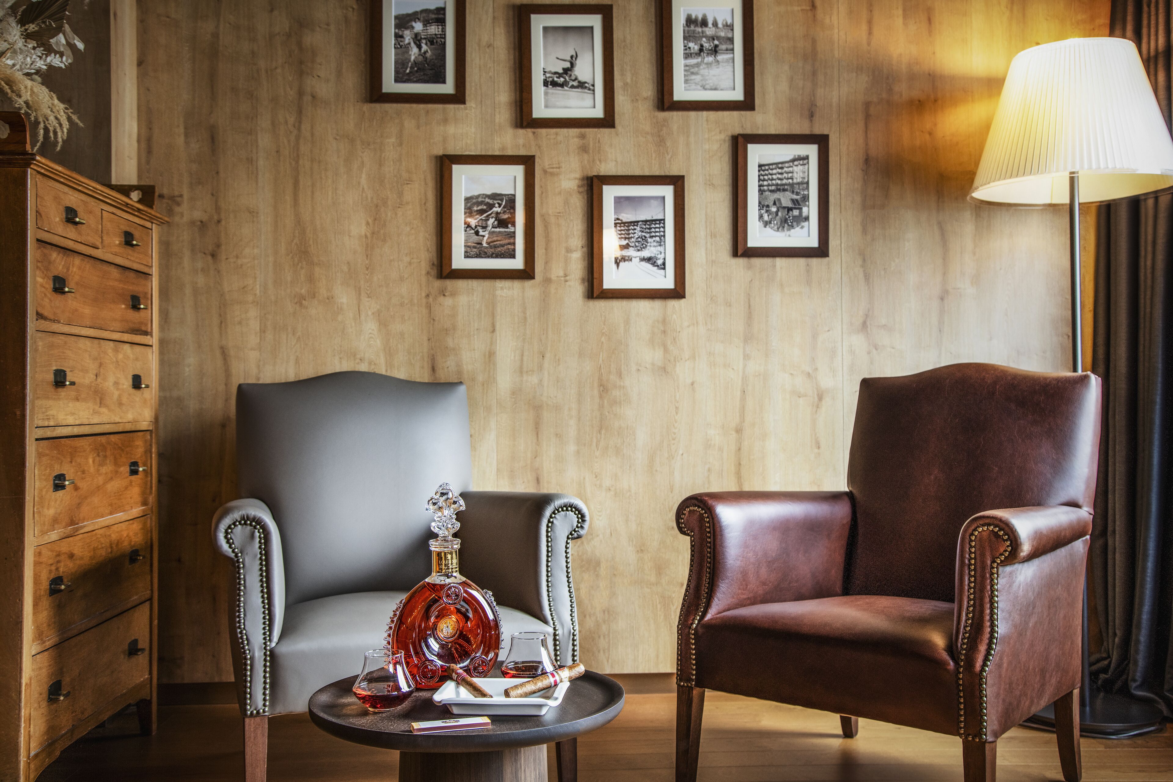 Una habitación con dos sillones de cuero, un tocador de madera, una mesa redonda con una botella y un vaso, y cuadros enmarcados en la pared.