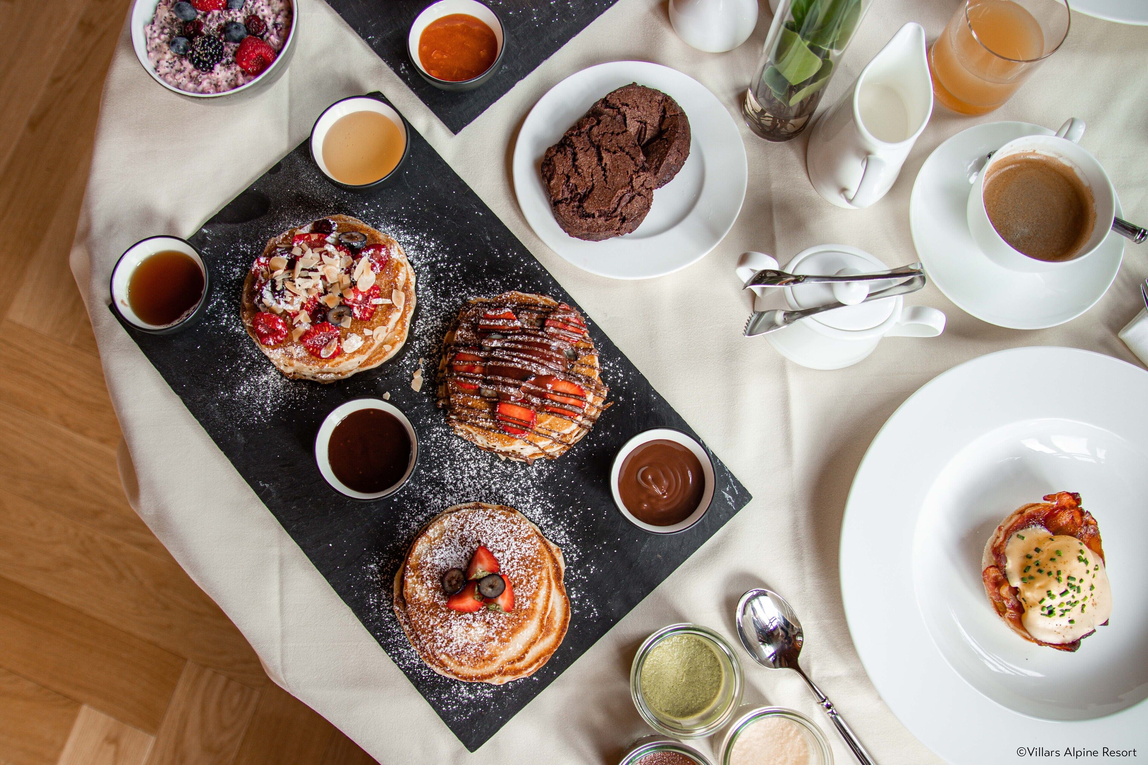 Ein Frühstücksarrangement mit Pfannkuchen, Waffeln und Gebäck auf einer Schieferplatte, dazu Tassen Kaffee und Tee.