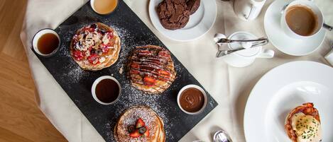 A breakfast setting with pancakes, waffles, and pastries on a slate board, accompanied by cups of coffee and tea.