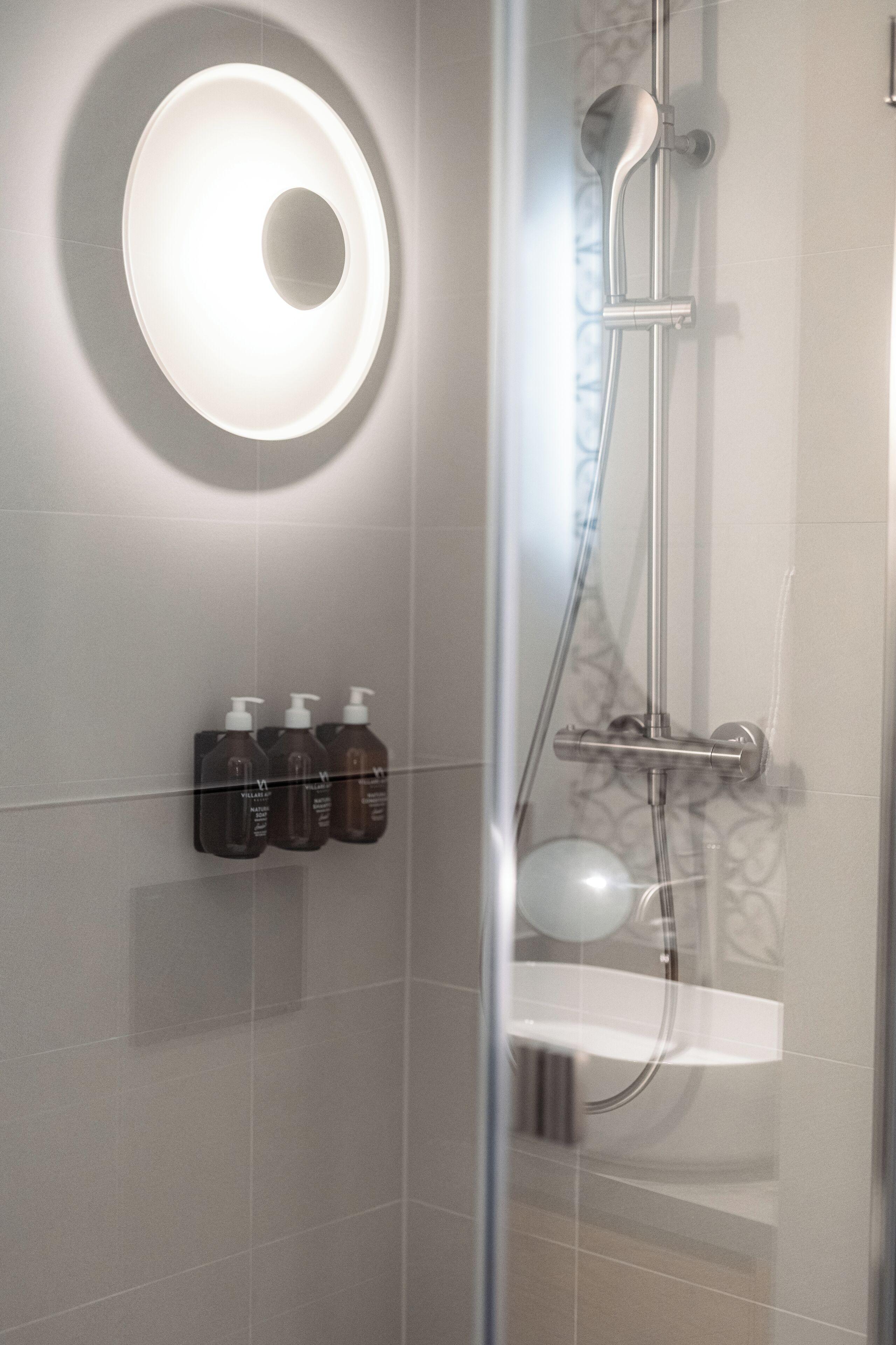 Ein modernes Badezimmer mit einer Glaskabine zur Dusche, einer Wandleuchte und drei Toilettenartikeln.