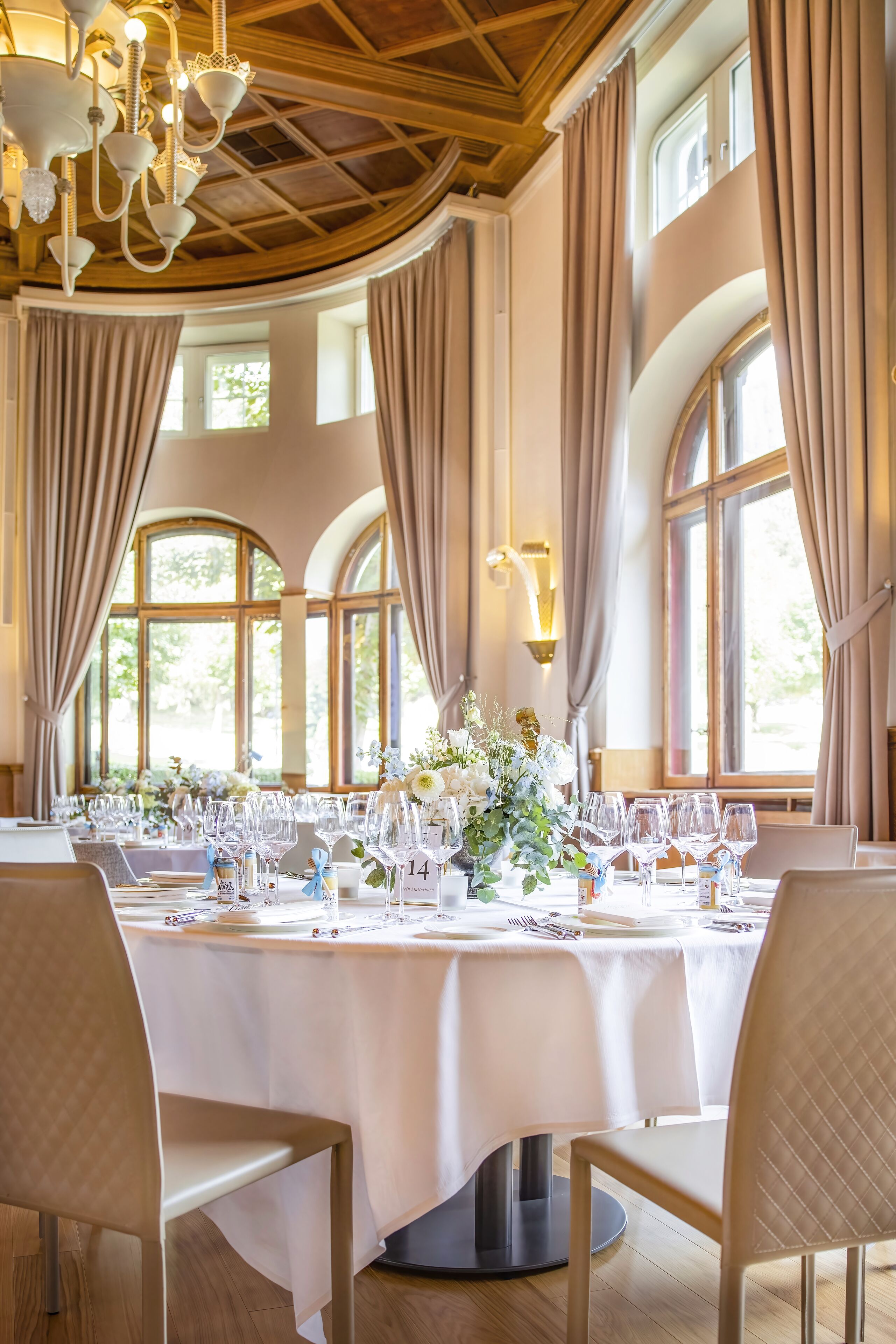 A dining area with round tables set for a meal, large windows with curtains, and a chandelier.