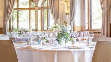 A dining area with round tables set for a meal, large windows with curtains, and a chandelier.