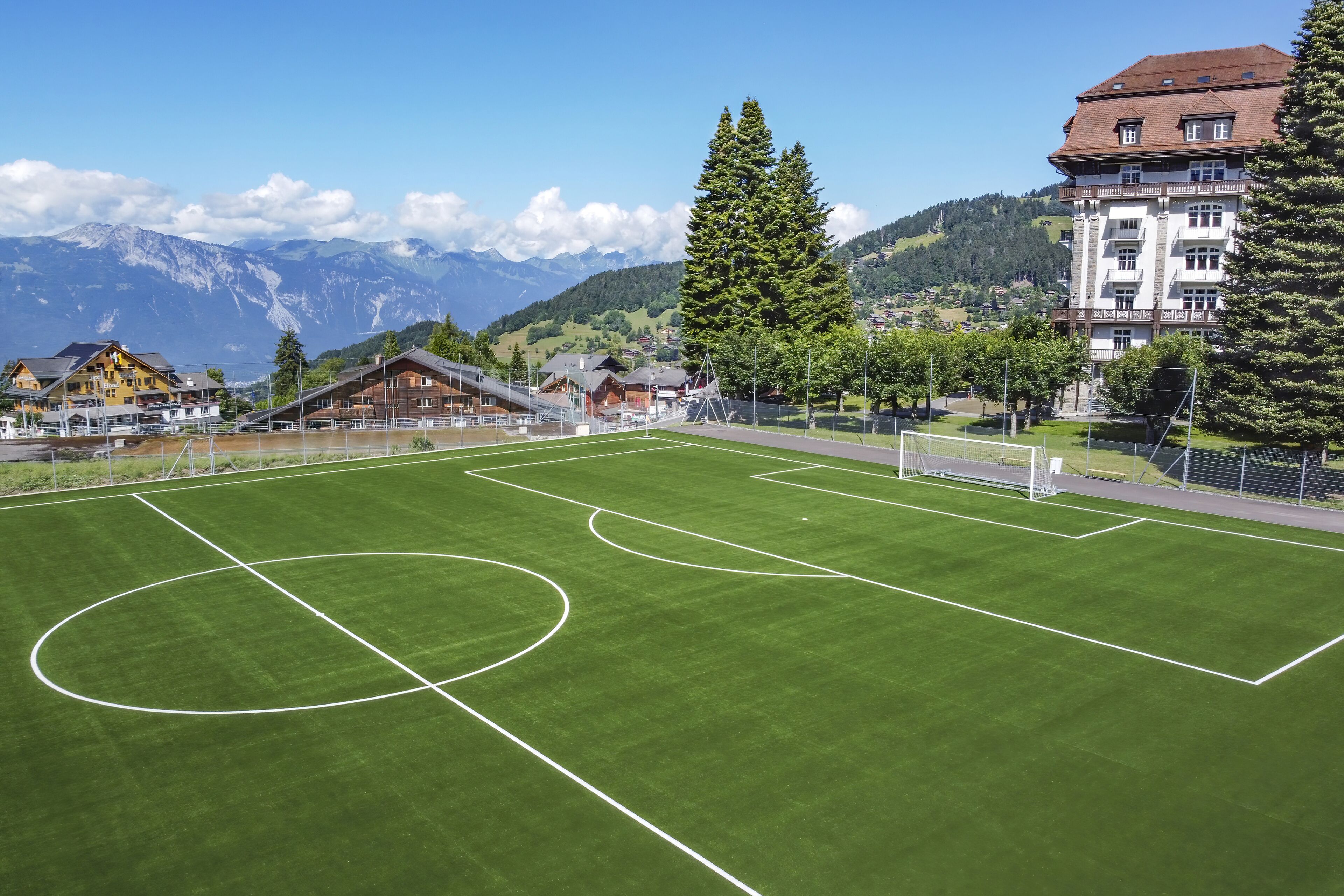 A soccer field with goalposts, surrounded by buildings and mountains in the background.