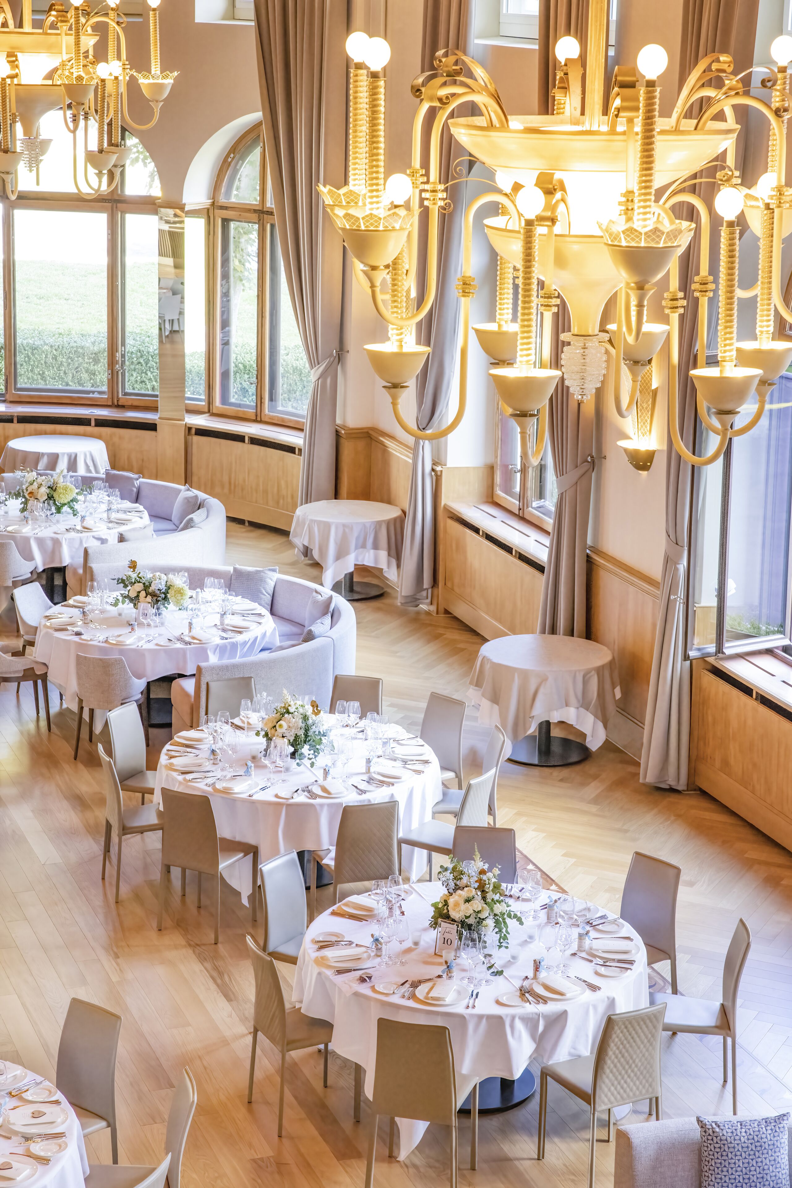 A dining area with round tables set for a meal, featuring chandeliers and large windows.