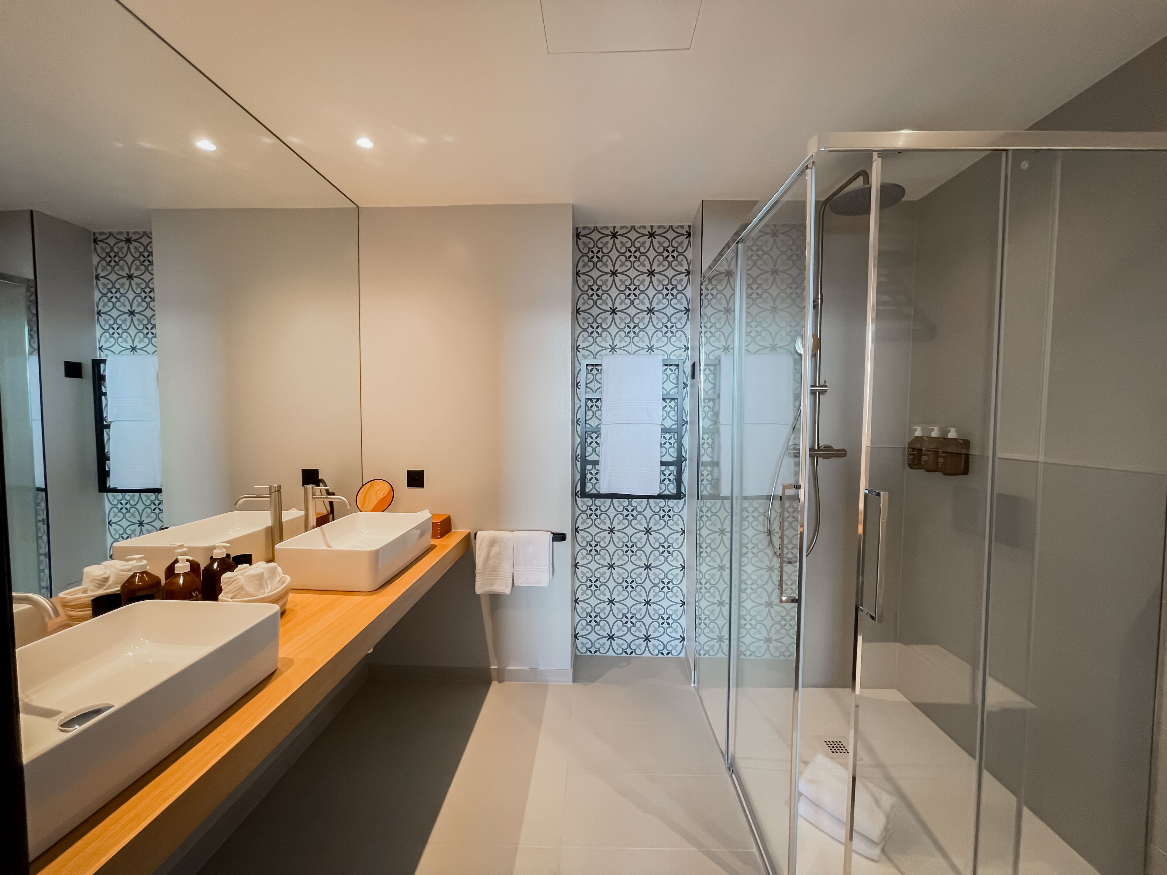 A modern bathroom with a large mirror, two sinks, a shower enclosure, and patterned wall tiles.