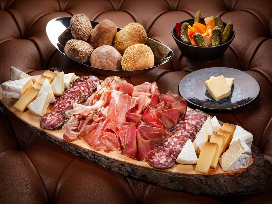 Una tabla de embutidos con diferentes carnes, quesos y pan sobre una tabla de madera, colocada sobre un sofá de cuero.