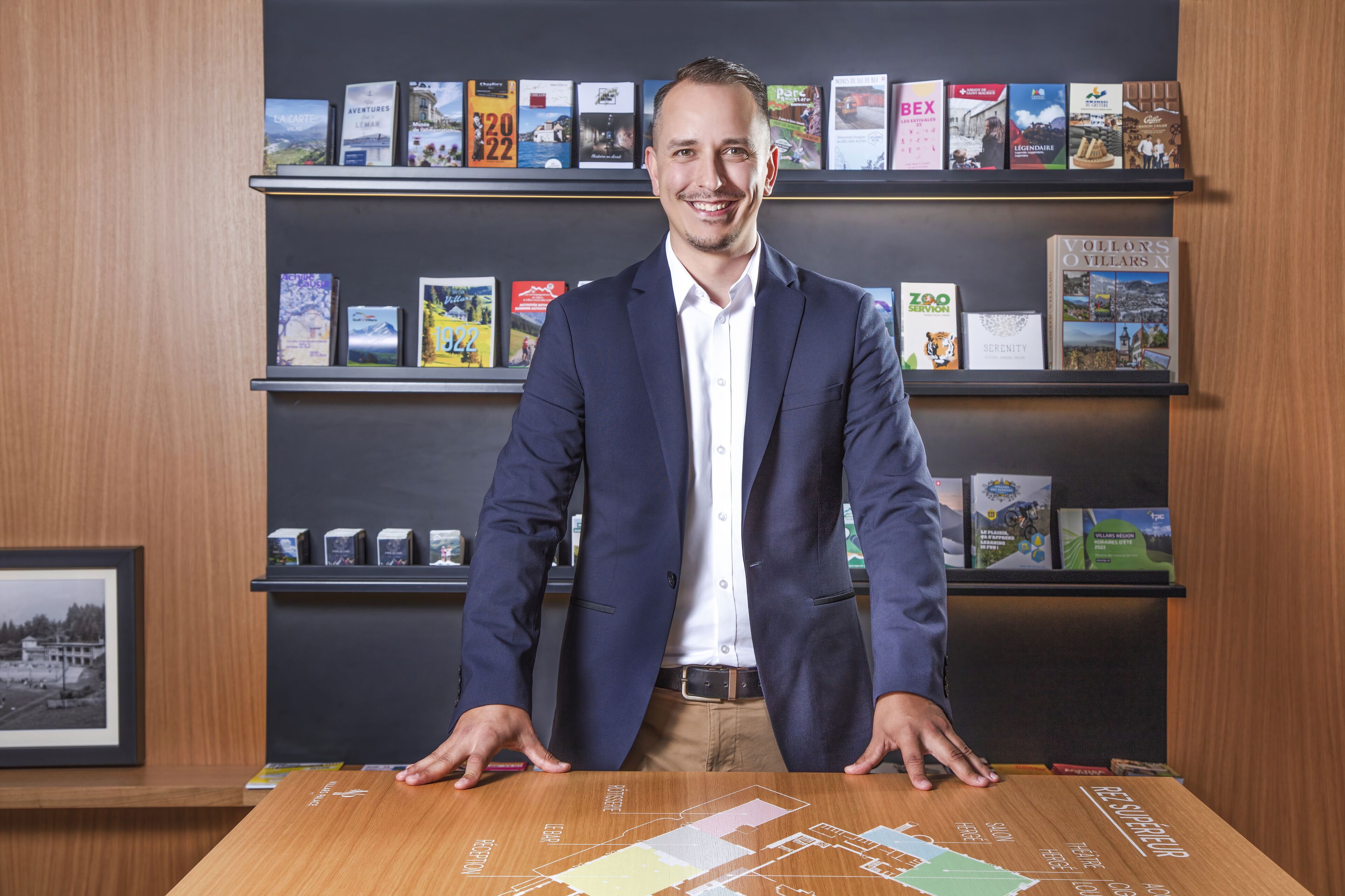 Un hombre con traje está de pie detrás de una mesa con un mapa, y detrás hay estanterías llenas de libros.