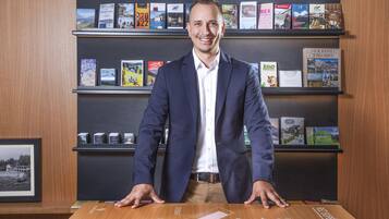Un hombre con traje está de pie detrás de una mesa con un mapa, y detrás hay estanterías llenas de libros.