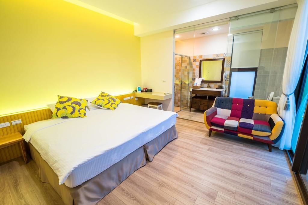 A hotel room with a large bed, a colorful sofa, a small desk, and a glass shower area.