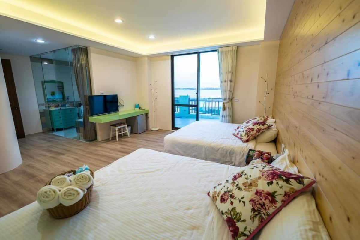 A hotel room with two beds, a desk, a television, and a view of the sea.