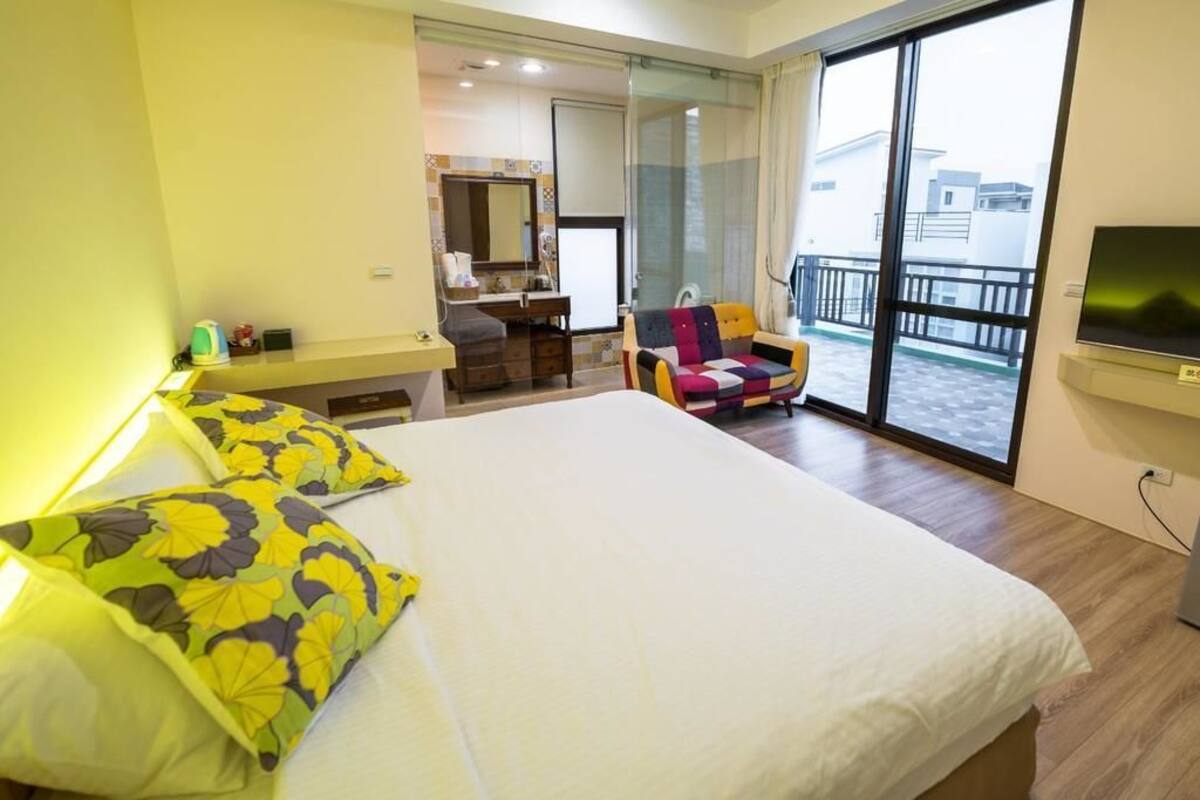 A hotel room with a large bed, a small desk, a chair, a TV, and a balcony with a view.