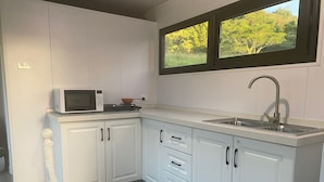 A compact kitchen with white cabinets, a microwave, a sink, and a window with a view of trees. - Nimbin Waterfall Retreat (Nimbin)