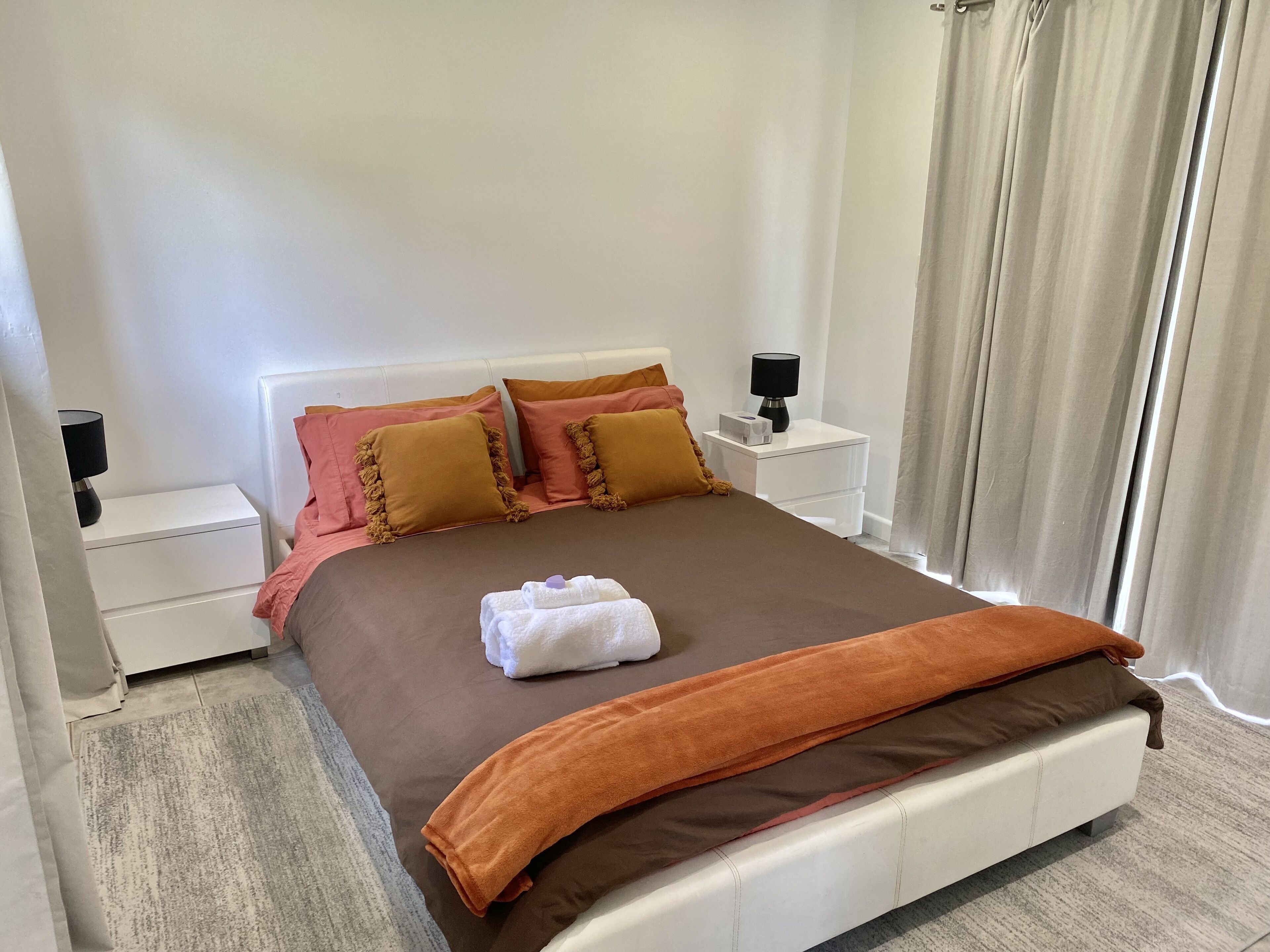 Ein ordentlich bezogenes Bett mit einer braunen und orangefarbenen Decke, zwei Nachttische mit Lampen und ein Fenster mit Vorhängen.