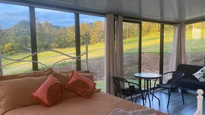 A bedroom with a large bed, a small table, two chairs, and a view of a green landscape. - Nimbin Waterfall Retreat (Nimbin)