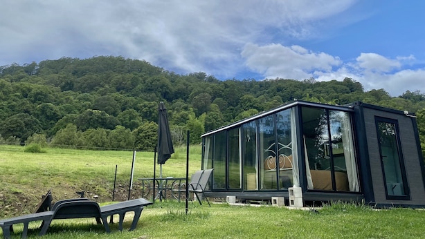 A modern glass house with a deck, surrounded by a grassy field and a forested hill. - Nimbin Waterfall Retreat (Nimbin)