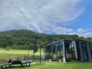 Una casa moderna de vidrio con terraza, rodeada por un campo cubierto de hierba y una colina boscosa.