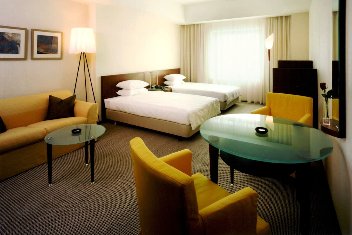 A hotel room with a bed, two bedside tables, a sofa, a chair, a glass table, and a lamp.