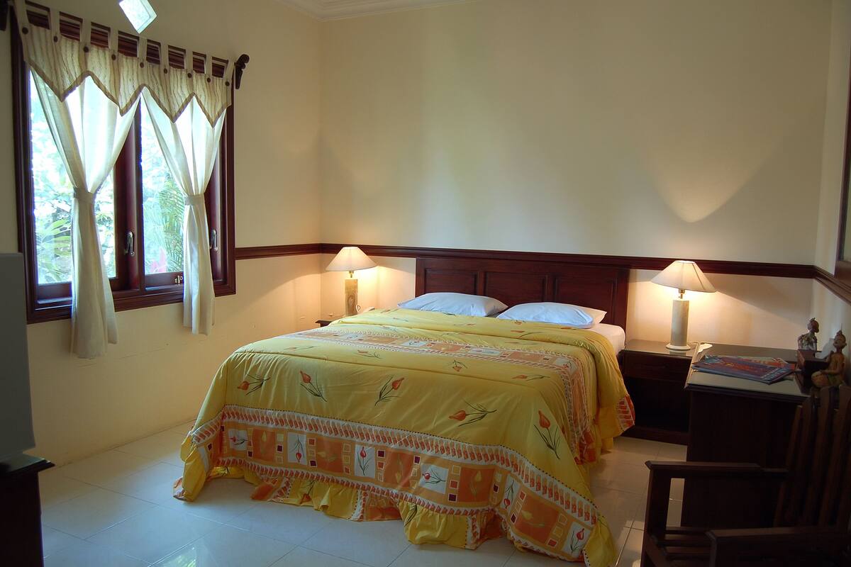 A bedroom with a bed, two bedside tables with lamps, a window with curtains, and a small table with a statue.