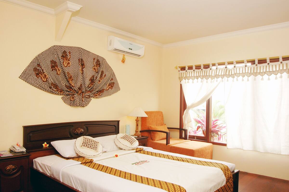 A hotel room with a bed, a chair, a nightstand, an air conditioning unit, and a window with curtains.