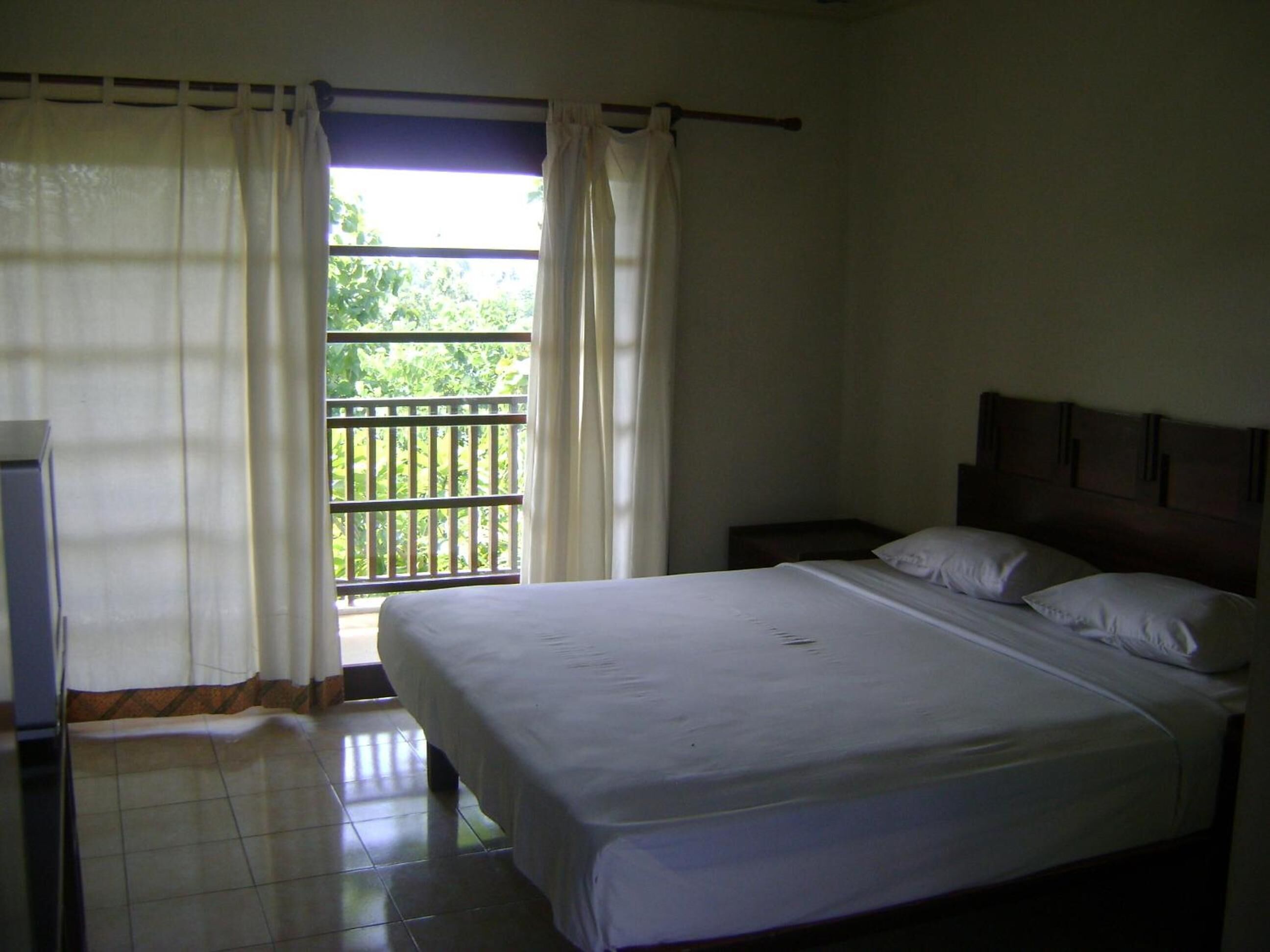 A bedroom with a bed, a nightstand, a window with curtains, and a view of greenery outside.
