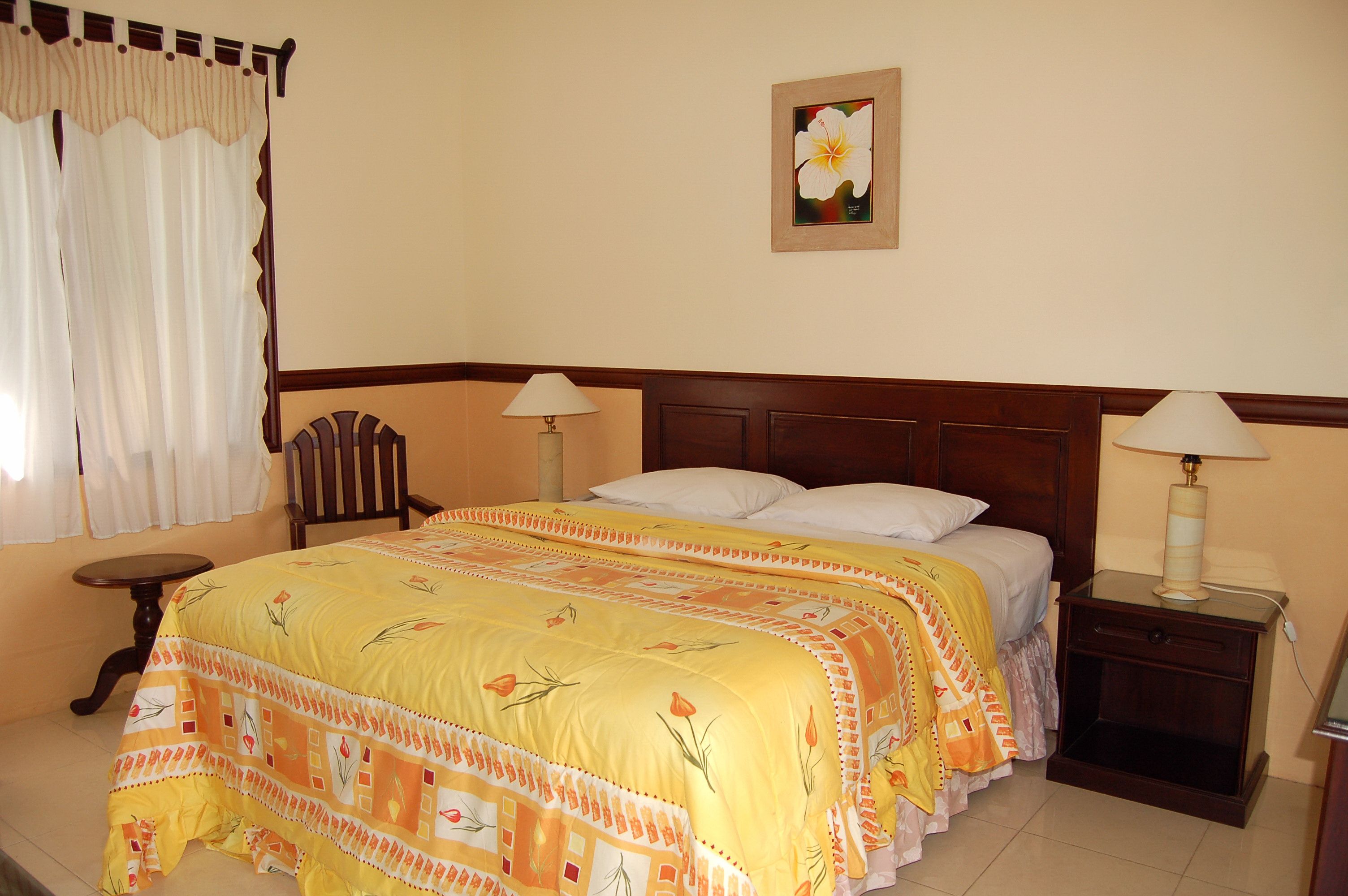 Una habitación de hotel con cama, dos mesitas de noche, una silla, un ventanal con cortinas y un cuadro colgado en la pared.