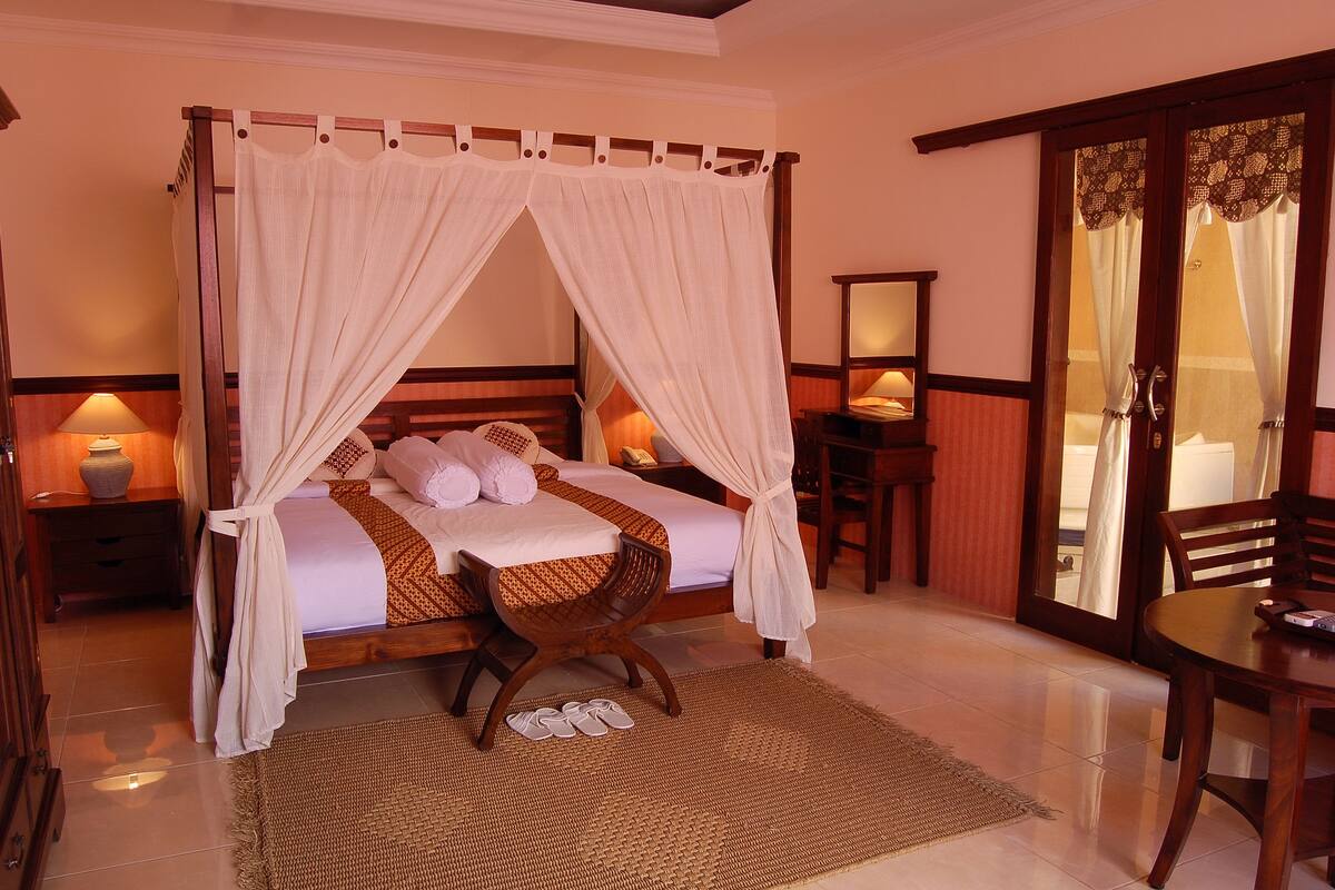 A four-poster canopy bed with a wooden headboard, a woven chair, and a small table with a lamp.