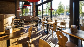 A spacious cafe with wooden tables and chairs, large windows offering a view of the street, and hanging pendant lights. - RiKu HOTEL Pfullendorf (Pfullendorf)