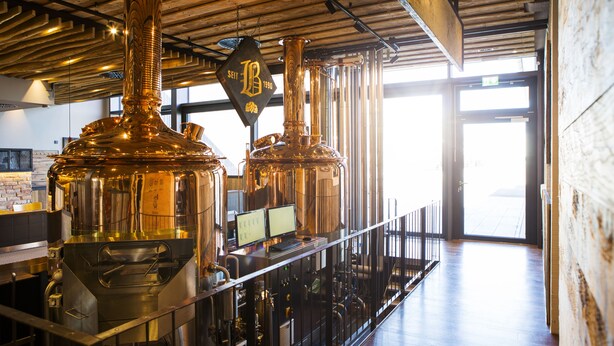 A brewery interior with stainless steel brewing equipment, a wooden ceiling, and a view of the outside through large windows. - RiKu HOTEL Pfullendorf (Pfullendorf)