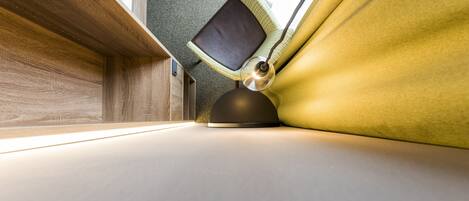 A modern, compact room with a wooden floor, a small table with a lamp, and a window providing natural light.