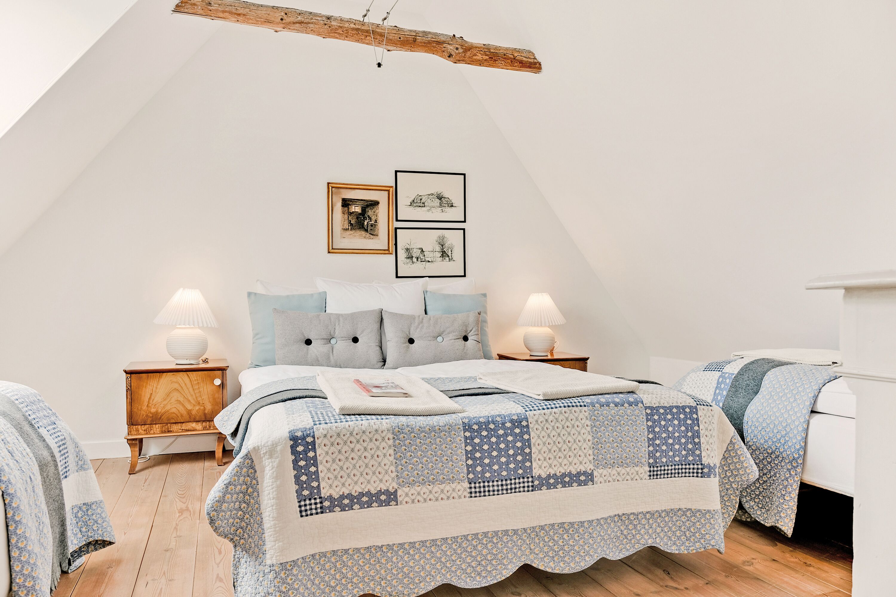 A bedroom with a sloped ceiling, a bed with a quilt, two bedside tables with lamps, and framed pictures on the wall.