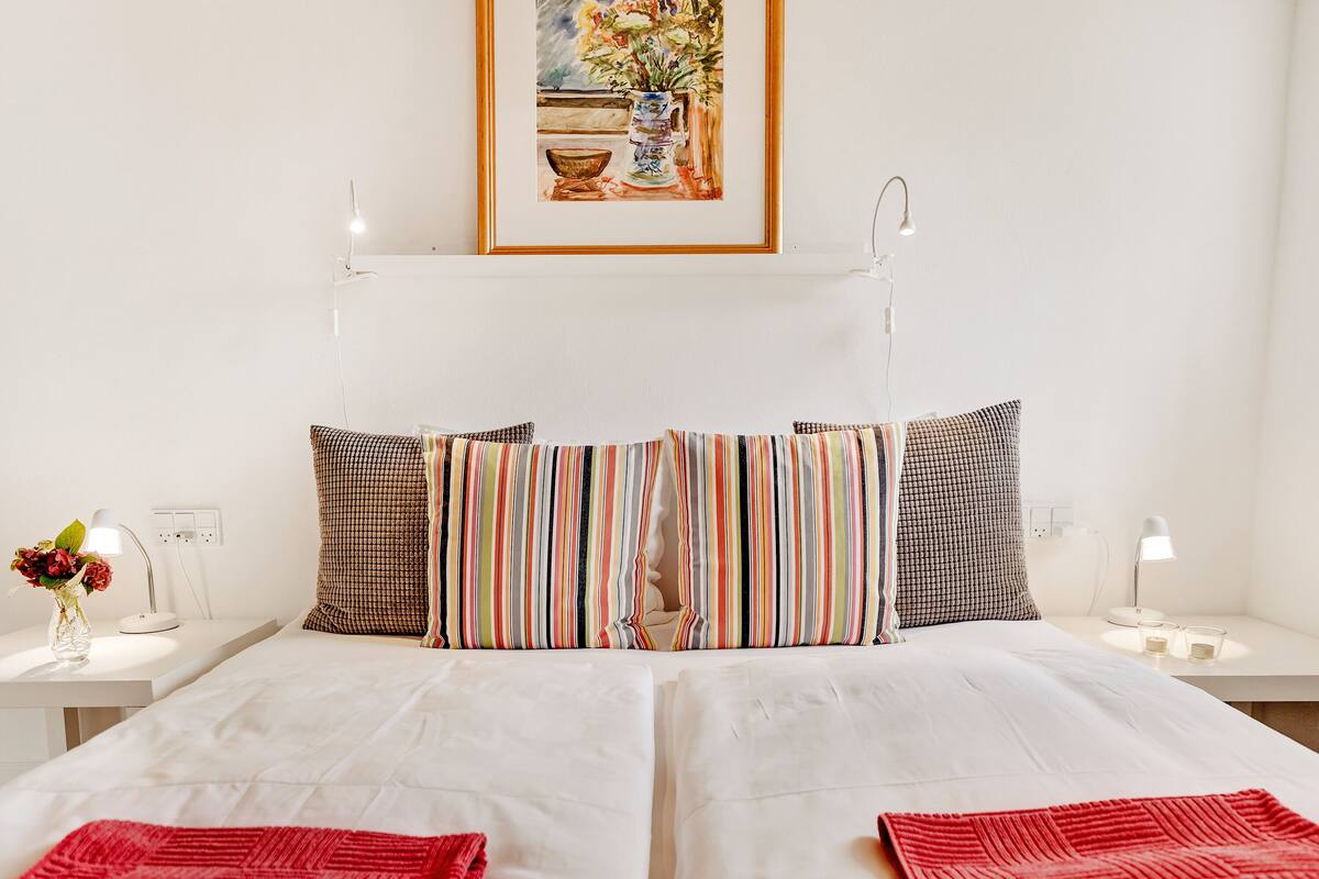 A hotel room with a bed, two bedside tables, a nightstand, a lamp, a vase with flowers, and a painting on the wall.
