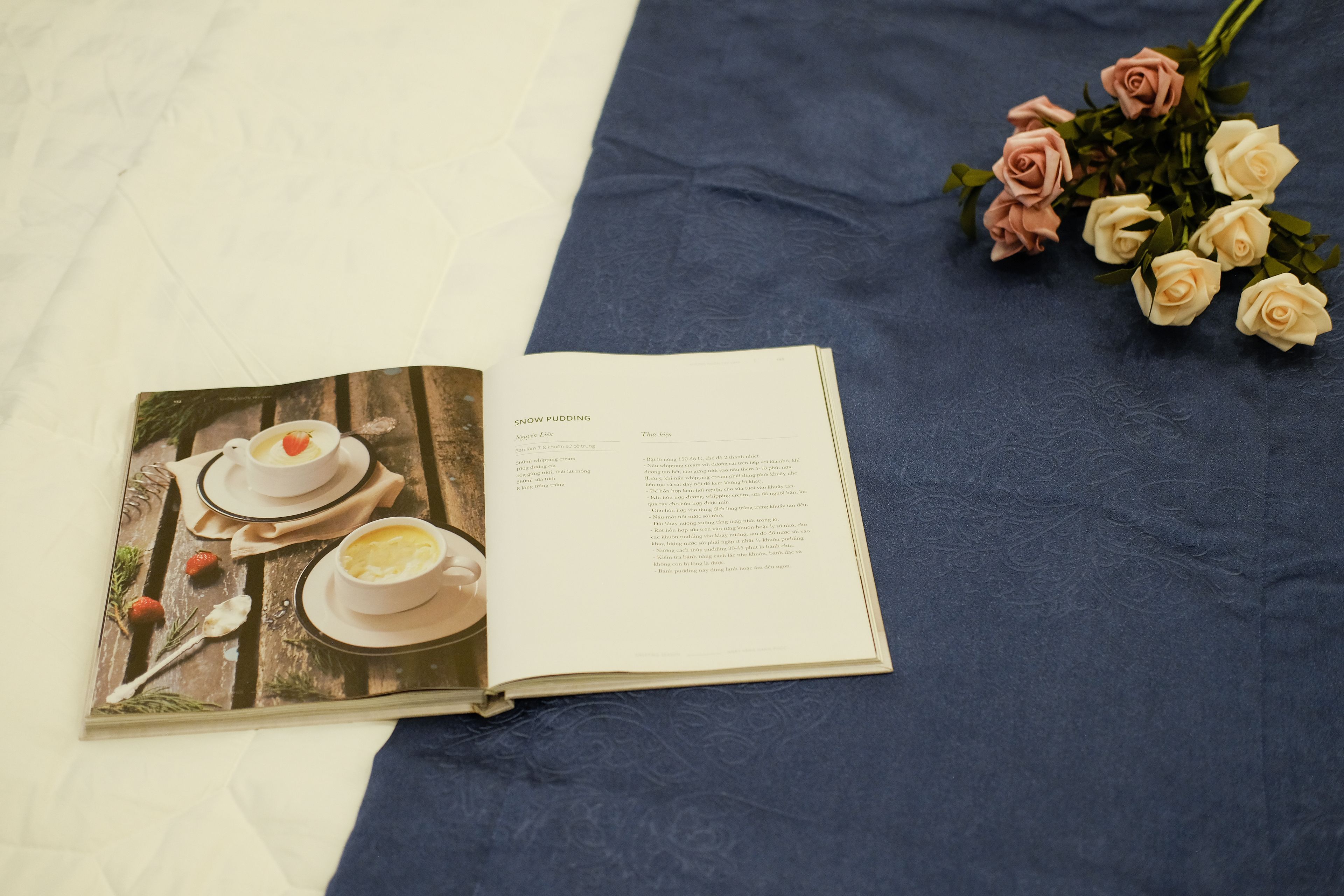An open book with a page displaying a dessert scene and a bouquet of roses on a blue fabric surface.