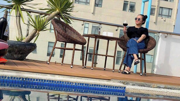 Une personne assise au bord d’une piscine, un verre à la main, entourée de meubles en osier et de plantes.