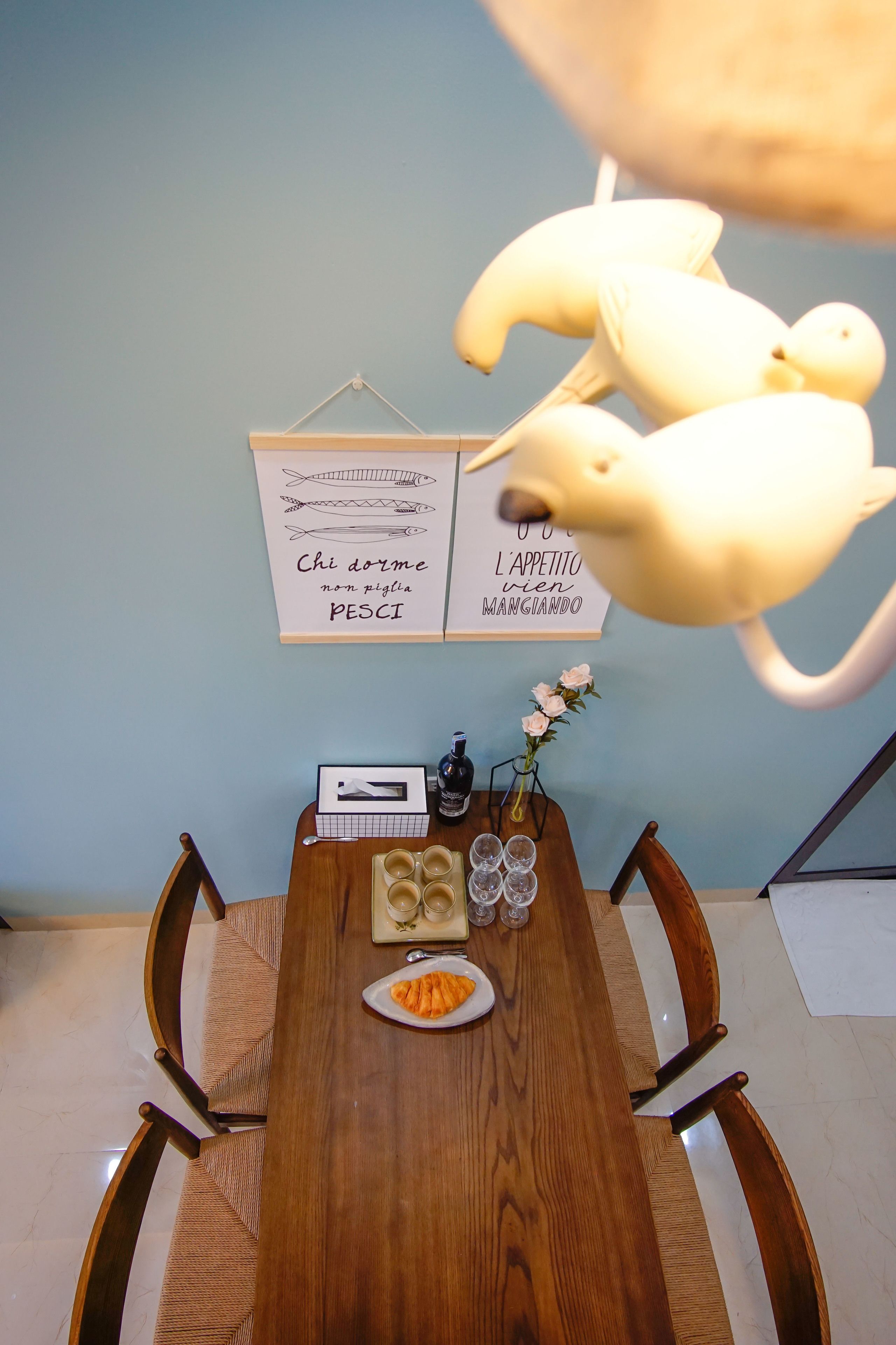 A dining table with chairs, a plate of food, a vase with flowers, a framed picture, and a hanging light fixture.