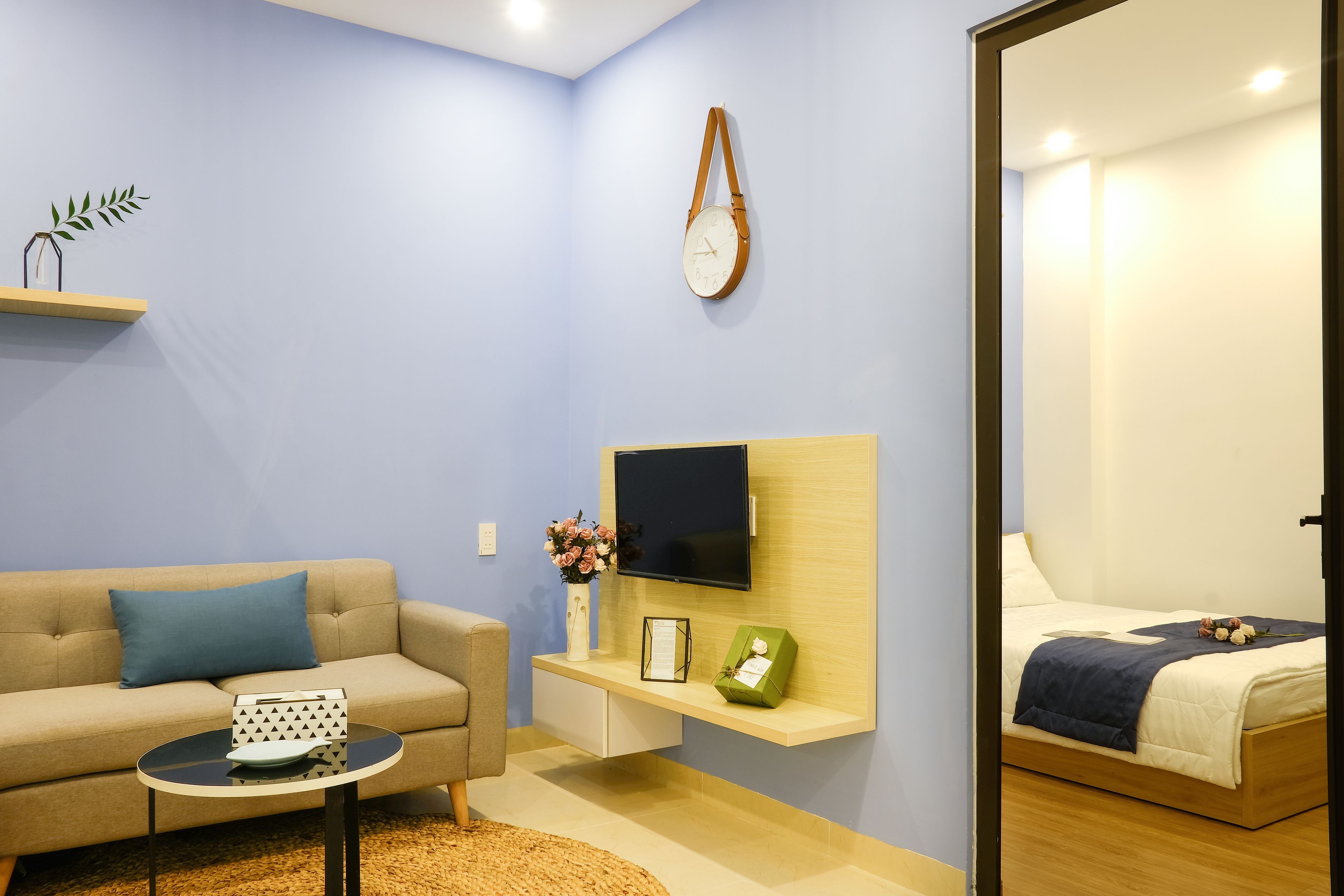 A modern living room with a sofa, a small round table, a mounted TV, and a bed with a blue blanket.