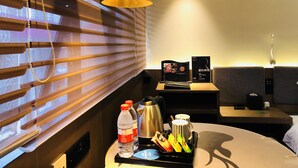 Habitación de hotel con cama, mesita de noche con lámpara, ventana con persianas, y una pequeña mesa con una bandeja que contiene una botella de agua, un termo y una taza.