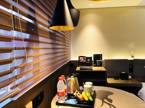 A hotel room with a bed, a bedside table with a lamp, a window with blinds, and a small table with a tray of items including a water bottle, a thermos, and a cup. - Likto Hotel (Guangzhou)