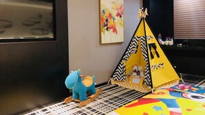 Una habitación con una pared negra, un cuadro abstracto y colorido, una carpa amarilla, un caballo balancín azul y un tapete estampado.