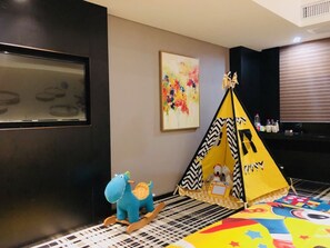 A room with a black wall, a colorful abstract painting, a yellow tent, a blue rocking horse, and a patterned rug. - Likto Hotel (Guangzhou)