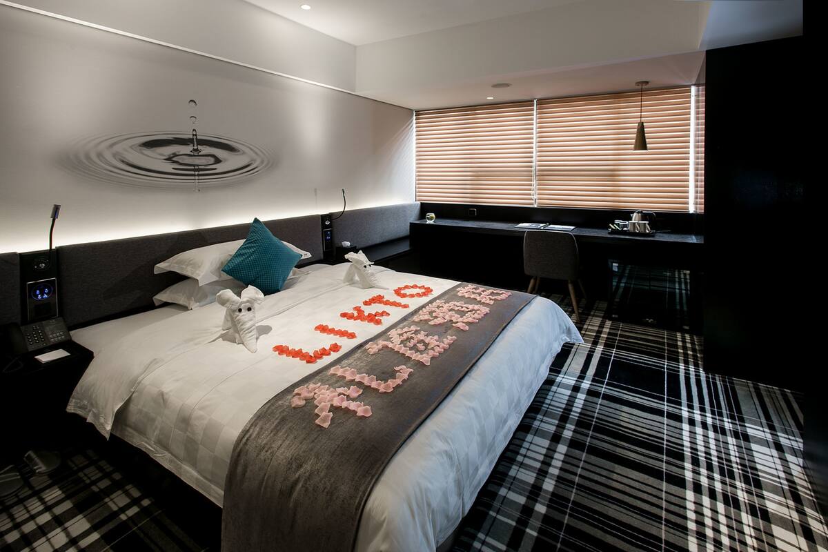 Ein modernes Hotelzimmer mit einem ordentlich bezogenen Bett, einem Nachttisch mit Lampe, einem Schreibtisch mit Stuhl und einem Fenster mit Jalousien.