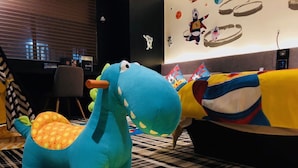 Una habitación con una silla con forma de dinosaurio azul, una manta amarilla y roja, y una pared con personajes de dibujos animados.