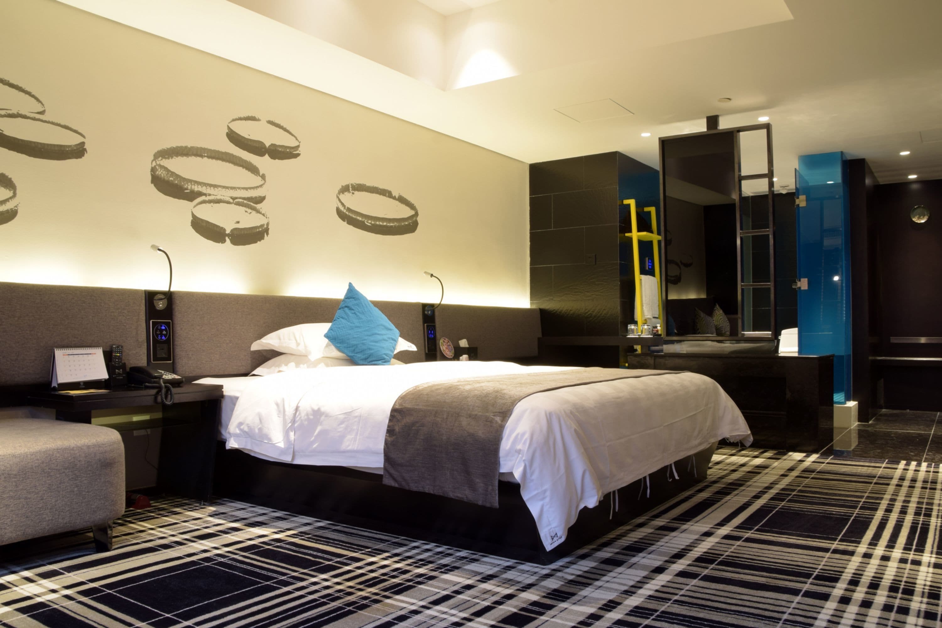 A modern hotel room with a large bed, a bedside table, a chair, a desk, and a mirror.