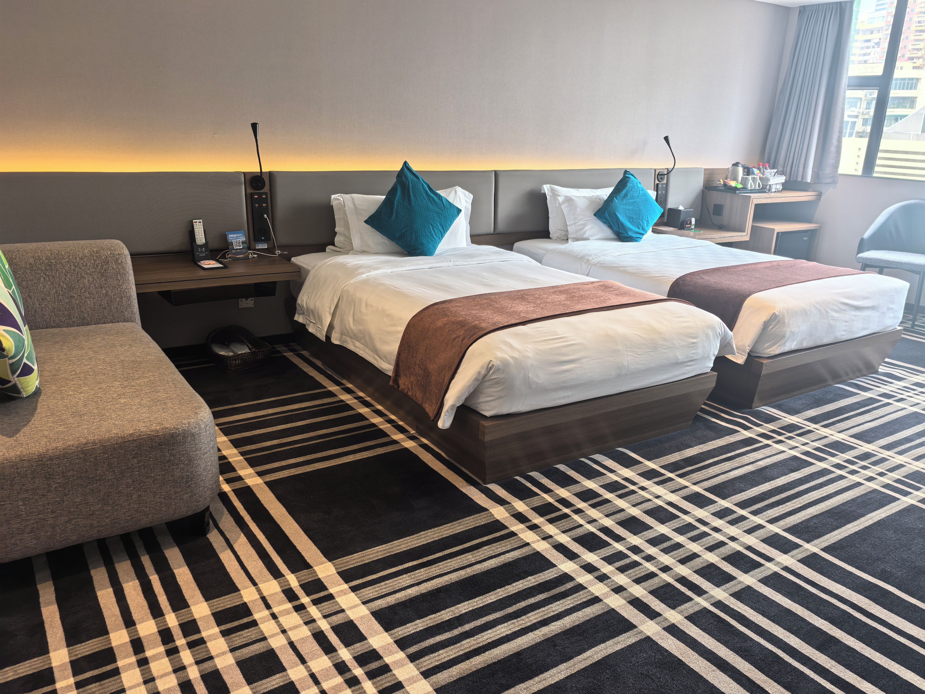 A hotel room with two beds, a sofa, a bedside table, and a large window.