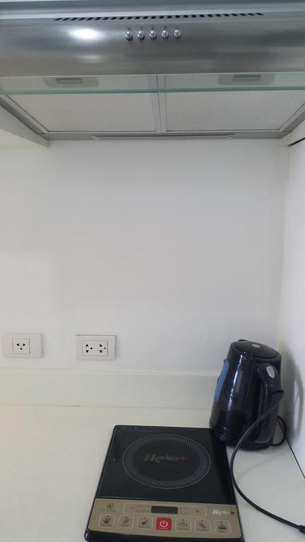 Una habitación con un hervidor eléctrico negro sobre una encimera, un enchufe y un estante de vidrio arriba.