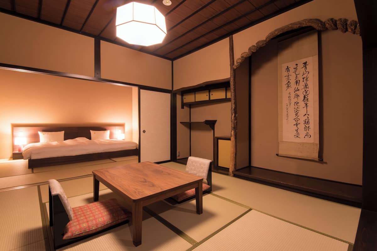 A traditional Japanese room with tatami flooring, a low wooden table, a cushioned bench, a bed with a headboard, and a wall with calligraphy.
