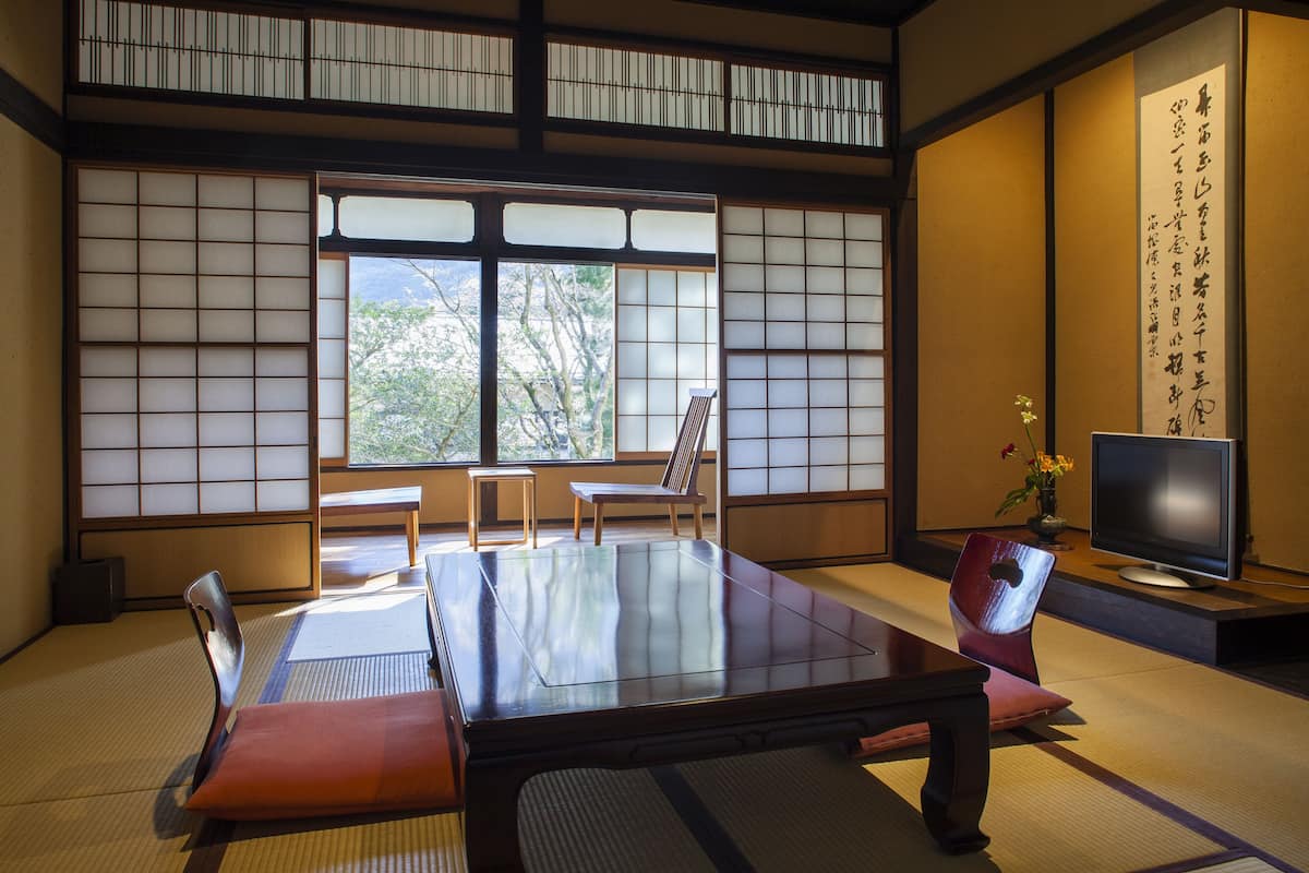 A traditional Japanese room with tatami flooring, a low table, two chairs, a television, and a large window with a view of trees.