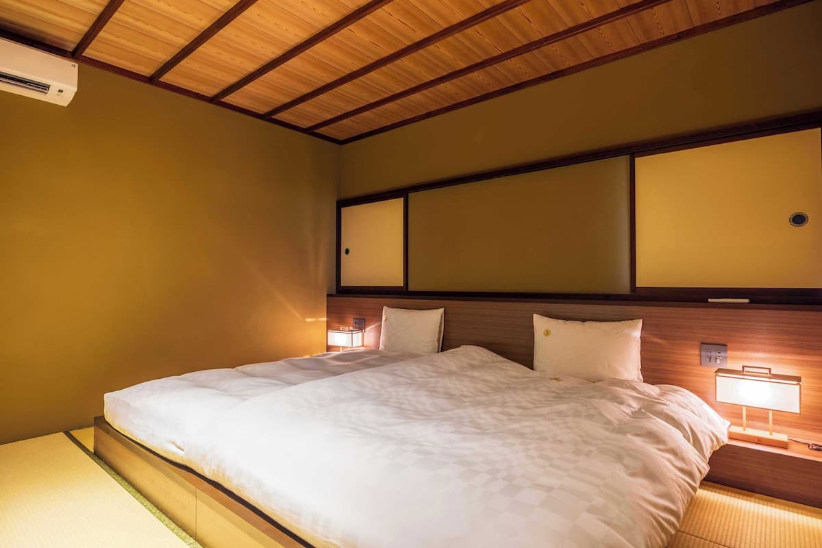 A hotel room with a wooden ceiling, a bed with white bedding, a nightstand with a lamp, and a wall-mounted air conditioner.