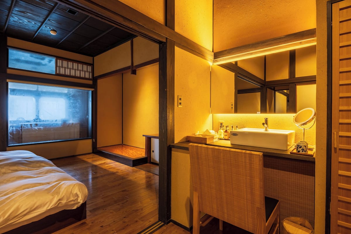 A hotel room with a bed, a small desk with a mirror, a sink, and a view through a large window.