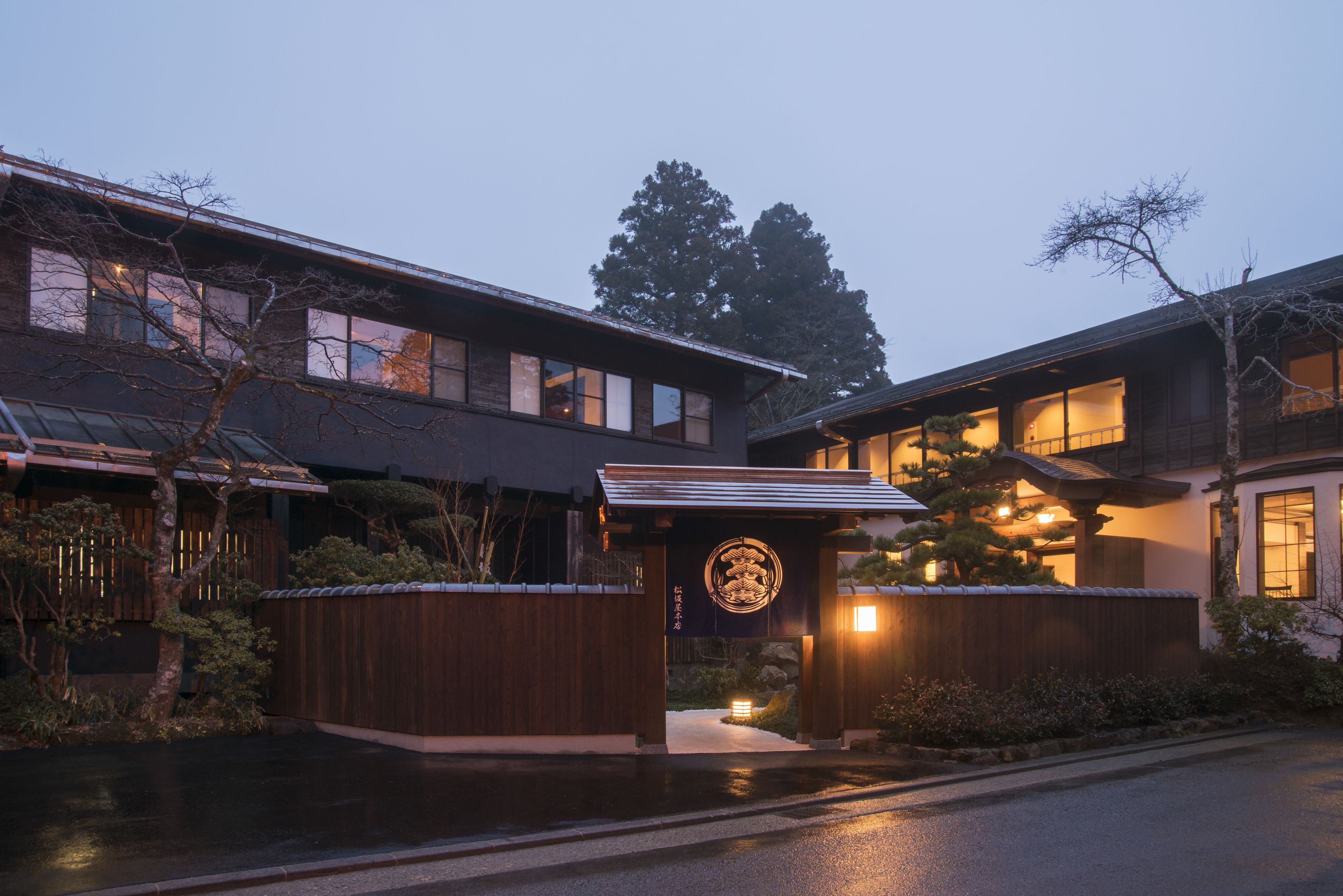A traditional Japanese building with a wooden entrance gate, a large tree, and a well-lit interior.