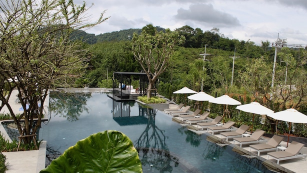 Una piscina infinita con sillones de descanso y sombrillas, rodeada de exuberante vegetación y un telón de fondo montañoso.